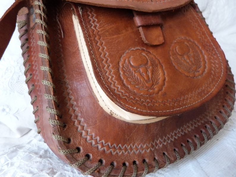 May include: A brown leather satchel bag with a flap closure and a decorative stitched design. The bag has two embossed circular designs with a stylized animal head and leaves.
