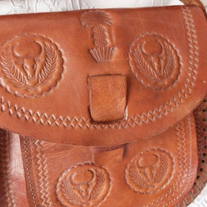 May include: A brown leather shoulder bag with intricate embossed designs of bull heads and a plant motif. The bag has a flap closure with a leather strap and a braided strap.
