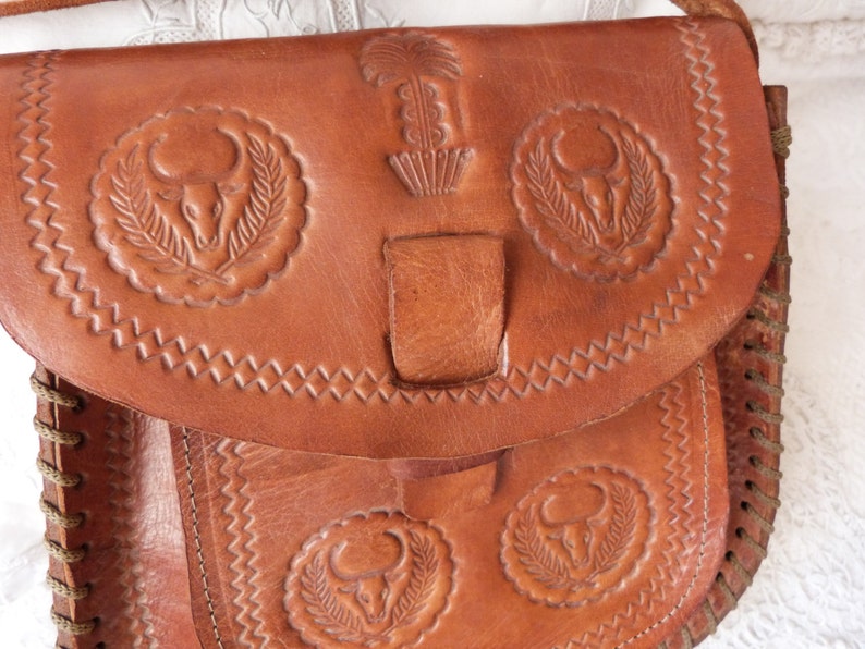 May include: A brown leather shoulder bag with intricate embossed designs of bull heads and a plant motif. The bag has a flap closure with a leather strap and a braided strap.