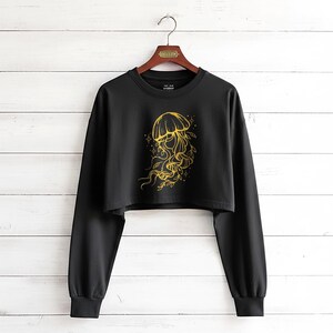 May include: Black cropped sweatshirt with long sleeves. The shirt features a gold illustration of a jellyfish with floral accents. The design is centered on the front of the shirt, creating a striking contrast against the black fabric.