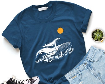 May include: Navy blue t-shirt featuring a white graphic of two whales and ocean waves under a yellow sun. The shirt is paired with blue jeans and black and white sneakers. A small succulent plant is also visible.