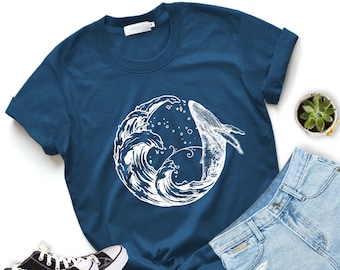 May include: A navy blue t-shirt featuring a white graphic of a whale breaching through ocean waves. The shirt is styled with rolled-up sleeves, a pair of light blue jeans, and black and white sneakers. A small succulent plant is also visible.