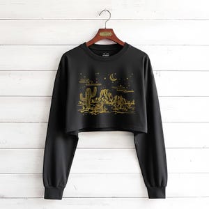 May include: Black cropped sweatshirt with a gold desert landscape design. The graphic features cacti, mountains, a crescent moon, and stars. The long-sleeved top is hanging on a wooden hanger.