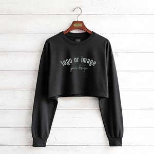 May include: Black cropped sweatshirt with long sleeves, hanging on a wooden hanger. The text "logo or image your design" is printed on the front in a cursive font. The background is a white wooden wall.