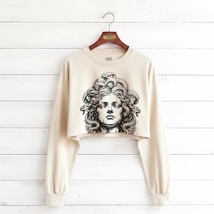 May include: A beige long-sleeve crop top featuring a black and white graphic print of Medusa with snakes for hair on the front. The garment has a crew neckline, dropped shoulders, and ribbed cuffs, displayed on a wooden hanger against a white background.