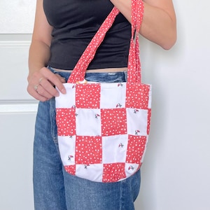May include: A tote bag featuring a checkered pattern of white squares and red squares with a white polka dot print. The bag includes two matching red and white polka dot fabric straps and small embroidered cherry motifs on the white squares.