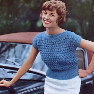 May include: A woman wearing a blue knitted sweater with short sleeves and a white skirt. She is smiling and standing next to a car.