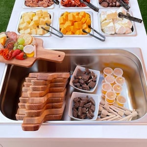 May include: A white buffet table with various food items arranged in stainless steel containers. The selection includes cheeses, meats, fruits, crackers, and chocolates. Wooden serving boards and honey sticks are also present, suggesting a catering or party setting.