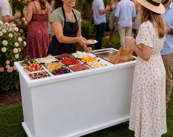 May include: A white mobile catering cart with a rectangular top surface featuring multiple stainless steel containers filled with assorted fruits, cheeses, meats, and crackers, positioned outdoors on a green lawn during a social gathering.
