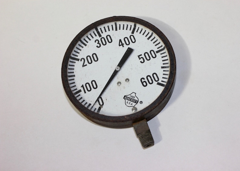 Pu&ograve; includere: Un manometro rotondo bianco con un ago nero che punta a 400. Il manometro ha un quadrante nero con numeri e segni bianchi. Il manometro &egrave; etichettato "1000 ASHCROFT U.S.A."