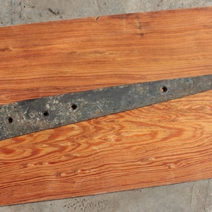 May include: A long, rusty, metal hinge with five holes and a pointed end. The hinge is lying on a wooden surface.