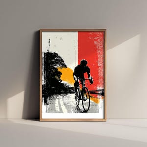 May include: A framed art print featuring a silhouette of a cyclist riding a road bike on a path. The background displays a split design with a white section on the left and a textured red section on the right, accented by a large yellow sun.