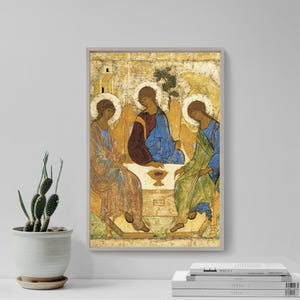 May include: Framed religious icon featuring three figures with halos, dressed in robes of blue, green, and brown. The artwork has a gold background and is set in a light-colored wooden frame. A small cactus in a white pot sits in front of the framed art.