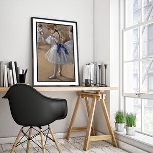 May include: A framed pastel drawing of a ballerina in a blue and white tutu, with other dancers in the background. The drawing is in a black frame and is hanging on a white wall above a wooden desk.