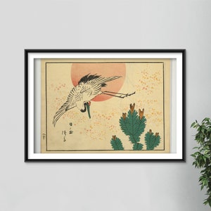 Op de afbeelding: Een vintage Japanse houtsnede die een witte kraan voorstelt die vliegt tegen een rode zon met groene dennentakken op de voorgrond. De print is zwart ingelijst en heeft Japanse tekst in de linkerbenedenhoek.