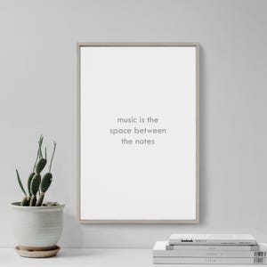 May include: A framed print with the text "music is the space between the notes" in a minimalist style. The print is in a light-colored frame and is displayed on a white surface. A small potted cactus and a stack of books are also visible.