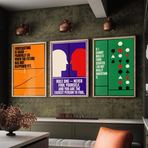 May include: Three framed wall art prints featuring graphic designs and text. The left print is orange with black text and a dotted line. The center print is purple with white and red silhouettes and text. The right print is green with text and shapes.