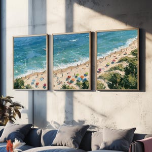 Op de afbeelding: Een drieluik van drie ingelijste wandkunstprints met een luchtfoto van een zandstrand met turquoise oceaangolven, verspreide kleurrijke parasols en kleine figuren van mensen die langs de kustlijn lopen, geplaatst in lichte houten lijsten.