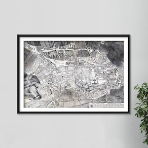 May include: Framed monochrome vintage map print. The detailed illustration depicts a town with streets, buildings, and fields. The print is in a black frame, mounted on a white wall. A green plant is visible in the corner.