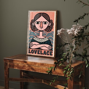 Op de afbeelding: Een rechthoekige ingelijste kunstdruk met een gestileerde illustratie van een persoon met donker haar in krullen, gekleed in een donkere jurk met een witte kraag. De achtergrond heeft blauwe golvende lijnen en roze spikkels. De tekst LOVELACE staat onderaan.