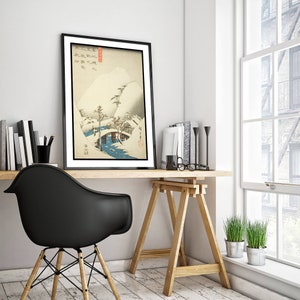 Op de afbeelding: Een zwart ingelijste print van een Japanse houtsnede die een besneeuwd landschap met een brug en bomen toont. De print hangt aan een witte muur boven een houten bureau met een zwarte stoel.