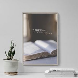 Può includere: Una stampa incorniciata con una citazione da Proverbi 31:25, "È vestita di forza e dignità: può ridere dei giorni a venire." La stampa presenta un primo piano di un libro aperto.