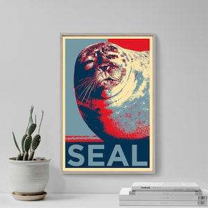 May include: Framed pop art print of a seal, using a limited palette of red, blue, and cream. The word "SEAL" is printed in bold, dark blue letters at the bottom of the image. The artwork is displayed in a neutral-toned frame.