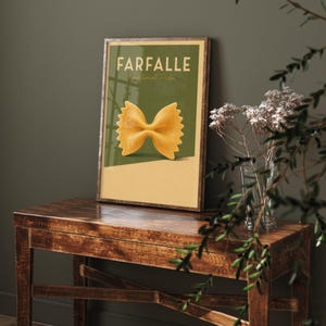 Pode incluir: Uma impressão de arte de parede retangular emoldurada apresentando uma grande forma de massa farfalle amarelo-dourada centralizada contra um fundo de dois tons de verde oliva e bege claro. O texto FARFALLE e Traditional Pasta está em branco.