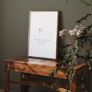 May include: A framed vertical wall art print featuring a white background with a small illustration of a person in a boat and the black text: Music washes away the dust of everyday life. — BERTHOLD AUERBACH. The frame has a thin, metallic finish.
