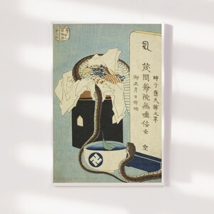 May include: A framed Japanese woodblock print depicting a snake coiled around a container, a bowl, and text. The artwork uses a blue and tan colour palette. The bowl has a symbol on it. The print is mounted on a white background.
