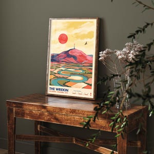 May include: A framed art print titled THE WREKIN WELLINGTON SHROPSHIRE 407M featuring a stylised landscape illustration with a red sun, a pink hill, a winding blue river, and a bird in flight against a yellow sky, with a scale bar at the bottom.