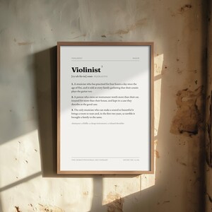 Könnte beinhalten: Ein gerahmter Kunstdruck mit einer wörterbuchartigen Definition des Wortes Violinist. Der Text enthält die phonetische Aussprache, eine Substantivbezeichnung und drei humorvolle nummerierte Definitionen. Der Druck befindet sich in einem hellen Holzrahmen.