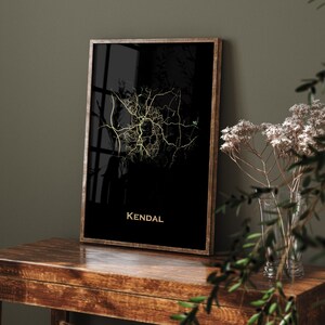 May include: A framed map print of Kendal, with a gold-colored street map design on a black background. The word "Kendal" is printed in gold at the bottom. The frame is a dark wood color. The print is displayed on a wooden table.
