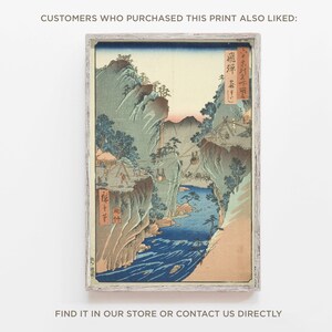 Op de afbeelding: Een vintage Japanse houtsnede die een berglandschap met een rivier die erdoorheen stroomt afbeeldt. De print is ingelijst in een wit houten lijst. De tekst in de afbeelding luidt "飛彈".