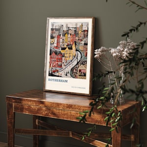 May include: A framed art print featuring a colorful illustration of Rotherham, South Yorkshire. The artwork depicts a street scene with buildings in shades of red, yellow, and brown. The print is displayed on a wooden table, with the text "ROTHERHAM" and "SOUTH YORKSHIRE" visible.