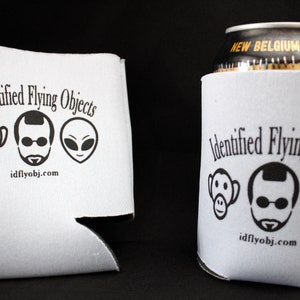 Może przedstawiać: Dwa białe piankowe ochładzacze do puszek z czarnym tekstem i grafiką. Tekst brzmi "Identified Flying Objects" i "idflyobj.com". Grafika przedstawia małpę, mężczyznę w okularach przeciwsłonecznych i kosmitę.