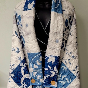 May include: A quilted wrap jacket featuring a blue and white floral pattern with a wide collar. The garment is constructed from patchwork fabric with visible stitching and includes two round wooden buttons at the lower front closure area.