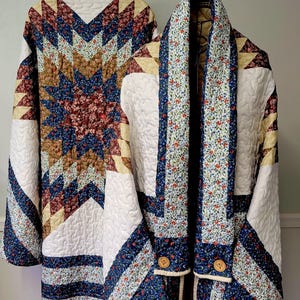 May include: A quilted jacket featuring a prominent star pattern in shades of navy blue, burgundy, and cream. The garment has a shawl collar, long sleeves, and two round wooden buttons at the front. The fabric displays a mix of floral and geometric prints.