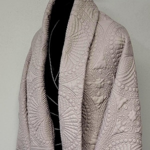 May include: A light beige quilted robe with intricate, circular embroidered patterns. The robe has a shawl collar and is displayed on a hanger. The robe's design features fan-like and floral motifs, creating a textured, elegant look.