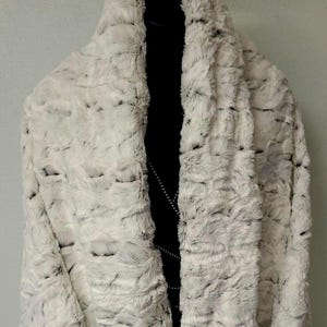May include: A plush, white faux fur wrap featuring a textured, mottled pattern with subtle black accents throughout the fabric. The garment has a wide, draped collar design and is displayed on a black mannequin, showcasing the thick, soft texture.