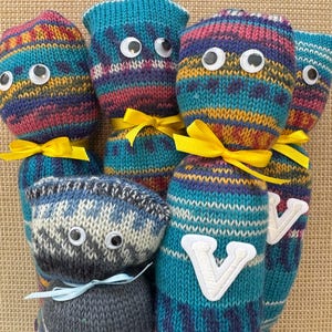 May include: A collection of whimsical, handmade knitted dolls with googly eyes. The dolls feature colorful striped patterns in shades of blue, teal, purple, and yellow. Some dolls have yellow or light blue ribbon bows, and some have a white embroidered "V" on them.