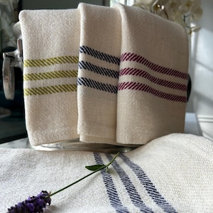 May include: Cream-coloured linen towels with horizontal stripes in olive green, navy blue, and burgundy. A stack of towels with yellow and navy blue stripes is in the foreground. A sprig of lavender rests on top.