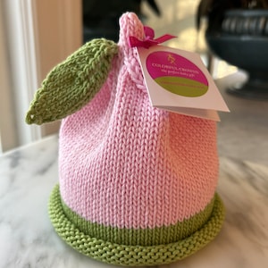 May include: A pink and green knitted hat shaped like a peach with a green leaf. The hat has a tag that reads "Colorful Crowns, the perfect baby gift, www.colorfulcrowns.com".