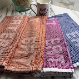 May include: Three folded kitchen towels featuring a herringbone weave pattern in orange, purple, and blue. Each towel displays the word EAT in large, light-colored block letters. The towels have white hemmed edges with thin blue and white striped accents.