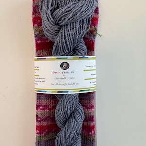 May include: A sock tube kit by Colorful Crowns. The kit includes a pair of patterned socks in shades of red, pink, and purple, along with a skein of gray yarn. The text on the packaging reads "You add the cuffs, heels, & toes."