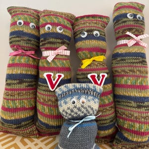 May include: A collection of handmade knitted cat dolls with googly eyes. The dolls are in various sizes and feature colorful striped patterns in shades of green, blue, and pink. Some dolls have bows and a letter "V" on them.