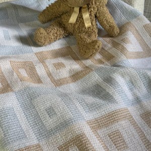 Peut inclure: Une couverture pour bébé tricotée et douce présentant un motif géométrique de carrés alternés bleu clair, blancs et beige. Un lapin en peluche texturé de couleur beige avec un petit ruban jaune clair noué autour du cou est posé dessus.