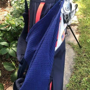 May include: A blue golf bag with a red and white strap. The bag has a white patch with a red, white, and blue American flag on it. The bag is hanging on a golf bag stand. The bag has the text "SUN MOUNTAIN® MISSOURI, MONTANA U.S.A." on it.