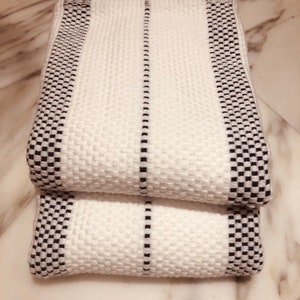 May include: Two folded white towels featuring a textured waffle weave pattern. The towels are accented with black and white checkered borders along the edges and a single thin black vertical stripe running down the center of each piece.