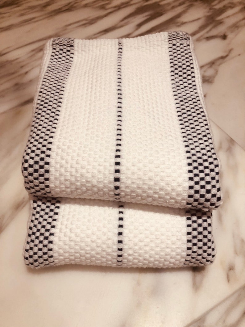 May include: Two folded white towels featuring a textured waffle weave pattern. The towels are accented with black and white checkered borders along the edges and a single thin black vertical stripe running down the center of each piece.